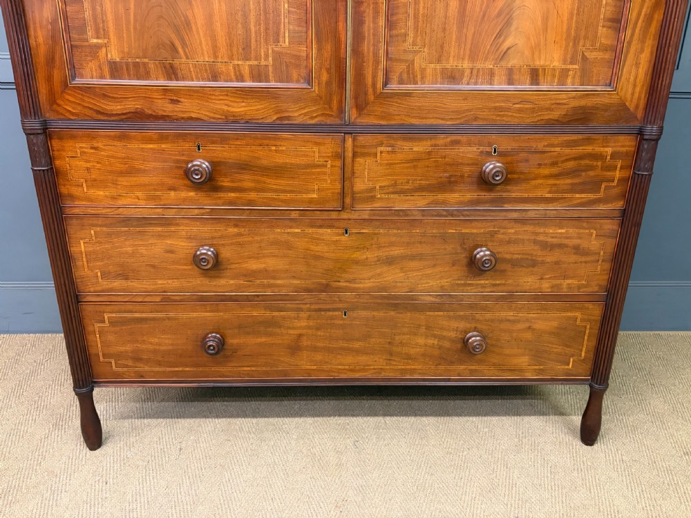 An antique Georgian flame mahogany linen press with an arched top cornice, two inlaid cupboard doors, fluted side pilasters, and a base of five drawers with turned knobs. View II