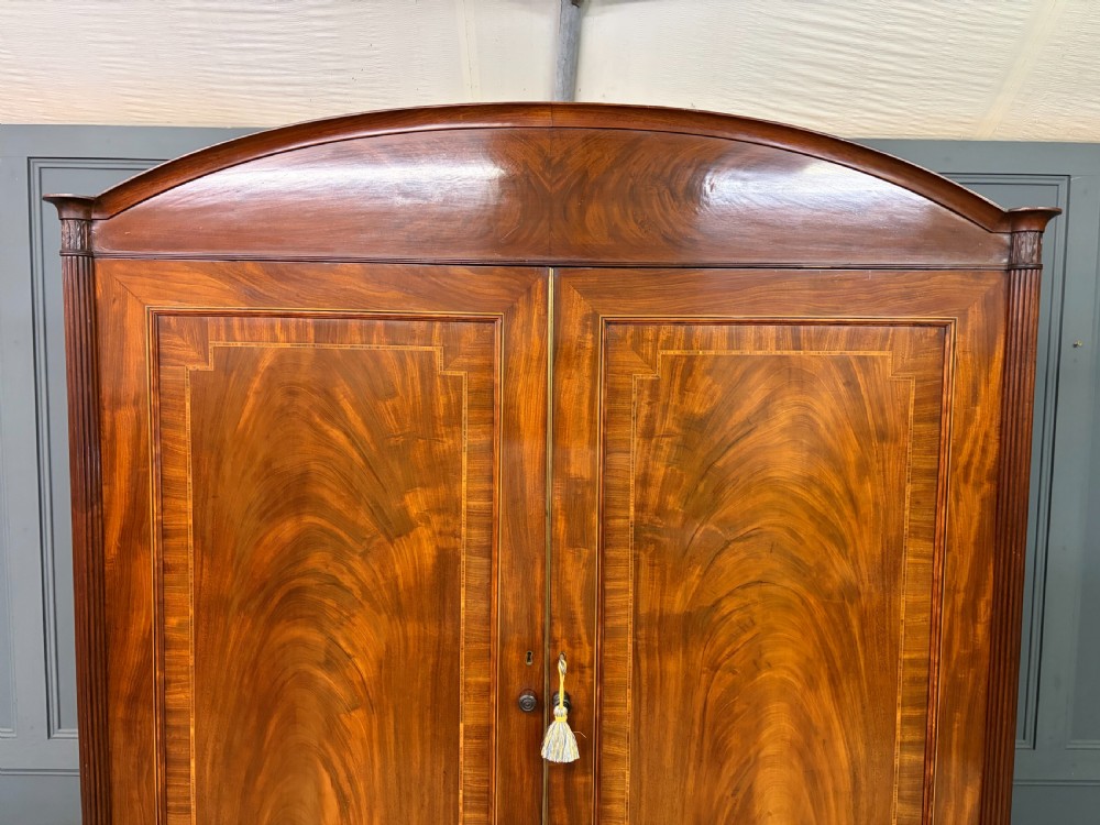 An antique Georgian flame mahogany linen press with an arched top cornice, two inlaid cupboard doors, fluted side pilasters, and a base of five drawers with turned knobs. View III