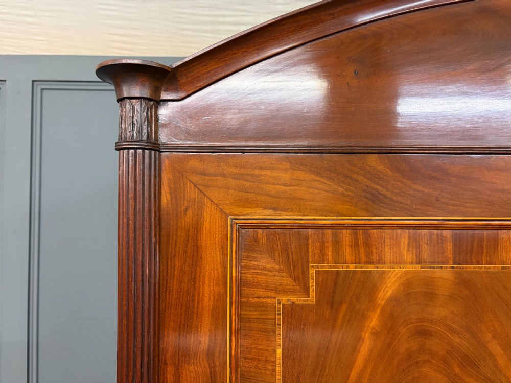 An antique Georgian flame mahogany linen press with an arched top cornice, two inlaid cupboard doors, fluted side pilasters, and a base of five drawers with turned knobs. View IV