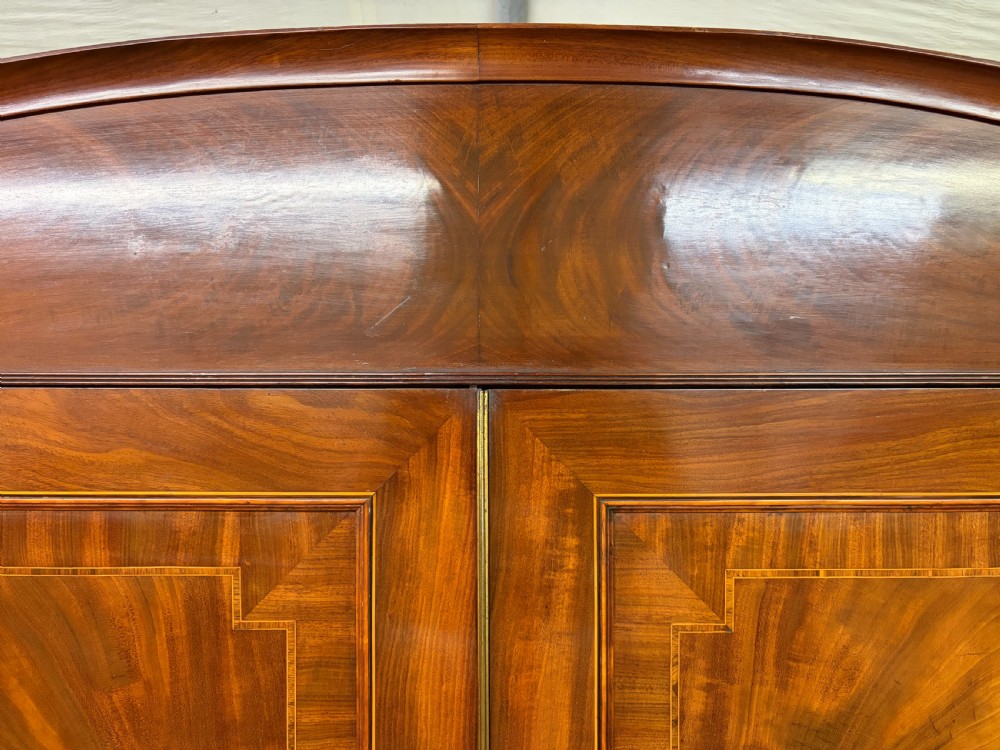 An antique Georgian flame mahogany linen press with an arched top cornice, two inlaid cupboard doors, fluted side pilasters, and a base of five drawers with turned knobs. View VI