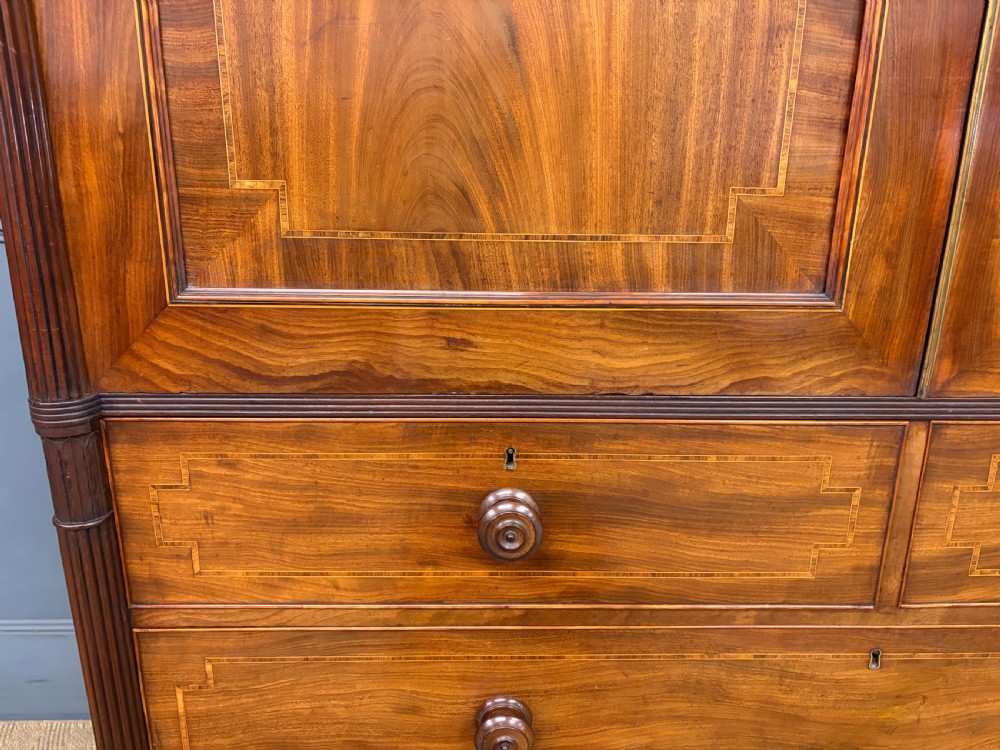 An antique Georgian flame mahogany linen press with an arched top cornice, two inlaid cupboard doors, fluted side pilasters, and a base of five drawers with turned knobs. View VII