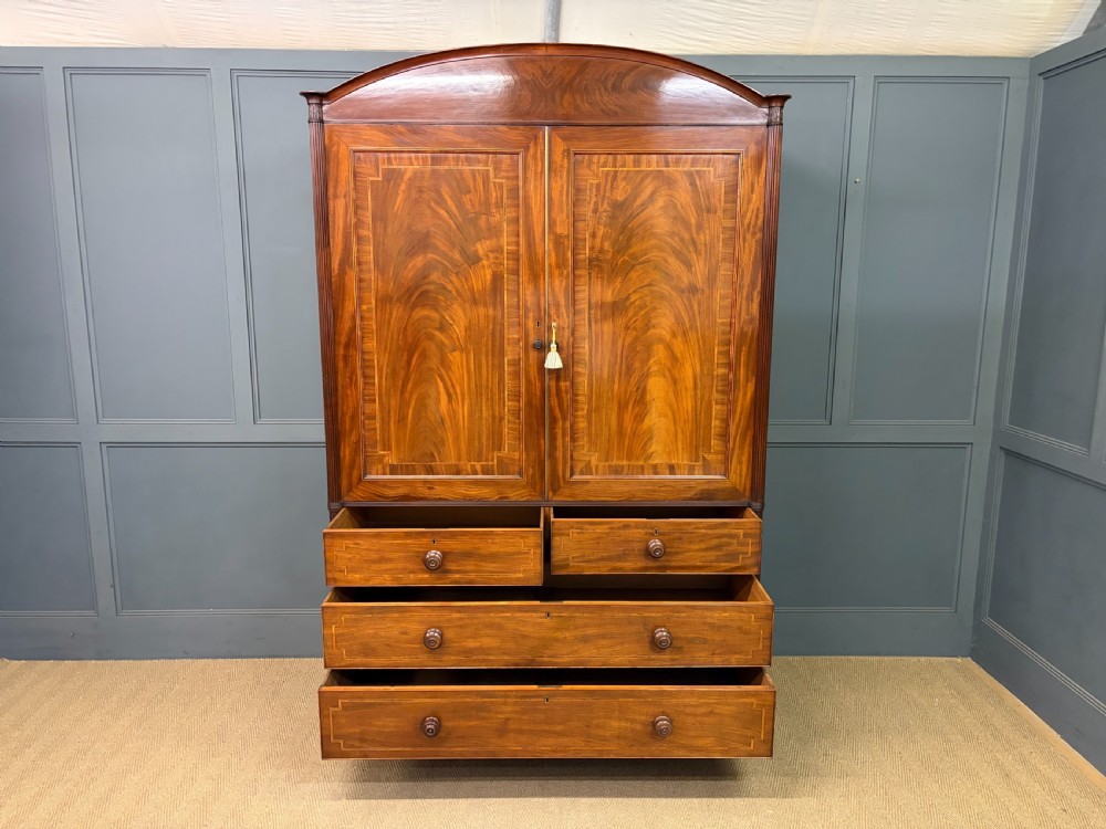 An antique Georgian flame mahogany linen press with an arched top cornice, two inlaid cupboard doors, fluted side pilasters, and a base of five drawers with turned knobs. View XIII