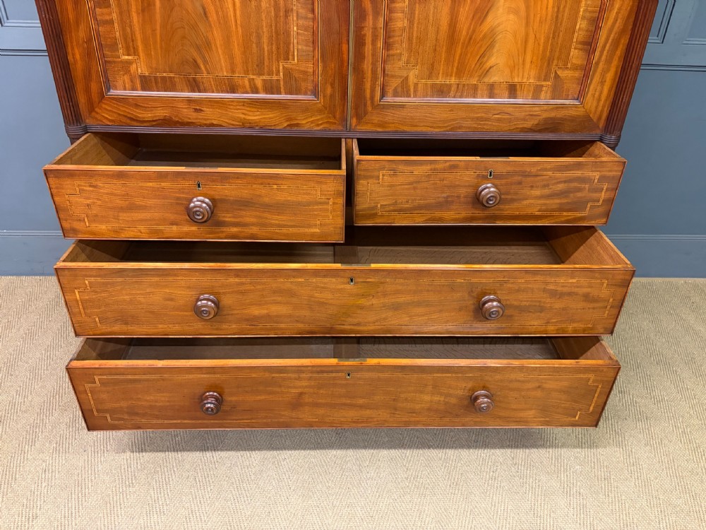 An antique Georgian flame mahogany linen press with an arched top cornice, two inlaid cupboard doors, fluted side pilasters, and a base of five drawers with turned knobs. View XIV