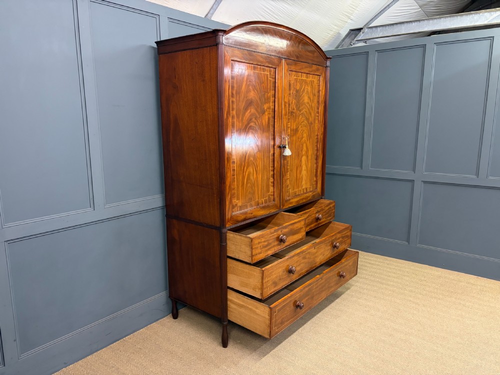 An antique Georgian flame mahogany linen press with an arched top cornice, two inlaid cupboard doors, fluted side pilasters, and a base of five drawers with turned knobs. View XV
