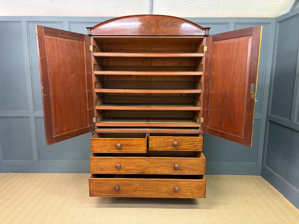 An antique Georgian flame mahogany linen press with an arched top cornice, two inlaid cupboard doors, fluted side pilasters, and a base of five drawers with turned knobs. View XVI