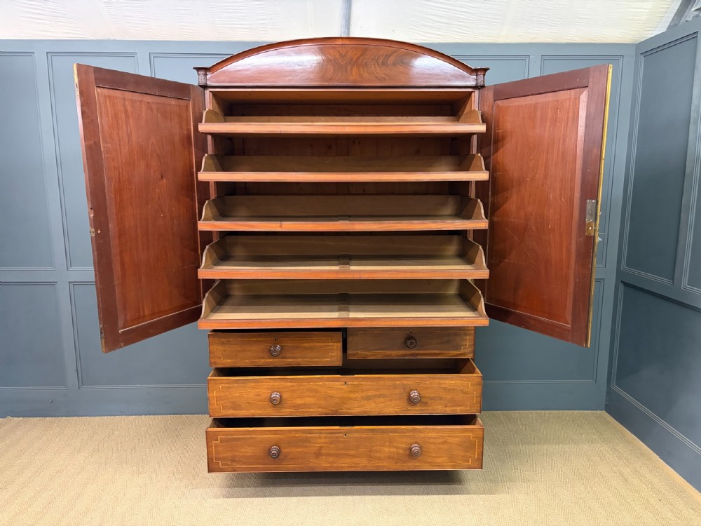 An antique Georgian flame mahogany linen press with an arched top cornice, two inlaid cupboard doors, fluted side pilasters, and a base of five drawers with turned knobs. View XVII