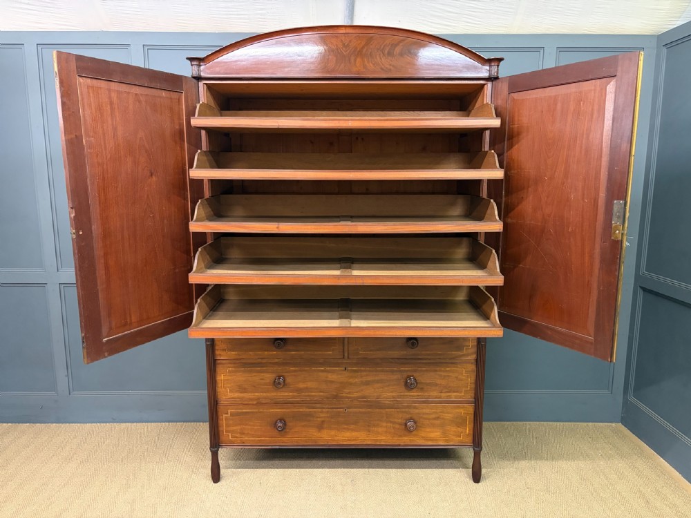 An antique Georgian flame mahogany linen press with an arched top cornice, two inlaid cupboard doors, fluted side pilasters, and a base of five drawers with turned knobs. View XVIII