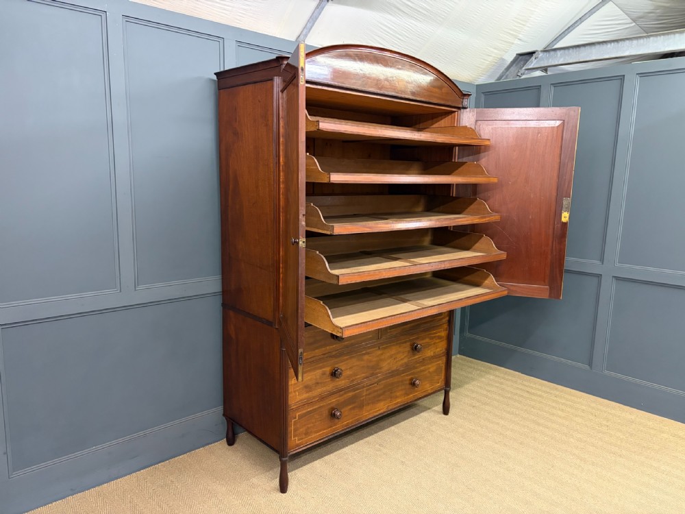 An antique Georgian flame mahogany linen press with an arched top cornice, two inlaid cupboard doors, fluted side pilasters, and a base of five drawers with turned knobs. View XIX
