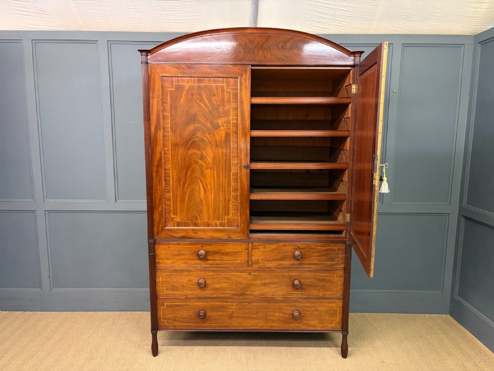 An antique Georgian flame mahogany linen press with an arched top cornice, two inlaid cupboard doors, fluted side pilasters, and a base of five drawers with turned knobs. View XXI