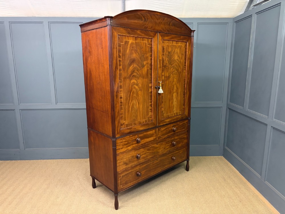 An antique Georgian flame mahogany linen press with an arched top cornice, two inlaid cupboard doors, fluted side pilasters, and a base of five drawers with turned knobs. View XXII