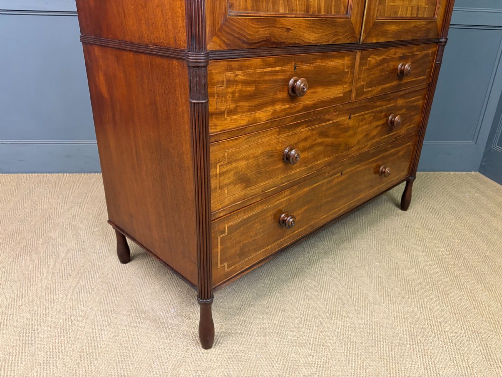 An antique Georgian flame mahogany linen press with an arched top cornice, two inlaid cupboard doors, fluted side pilasters, and a base of five drawers with turned knobs. View XXIII