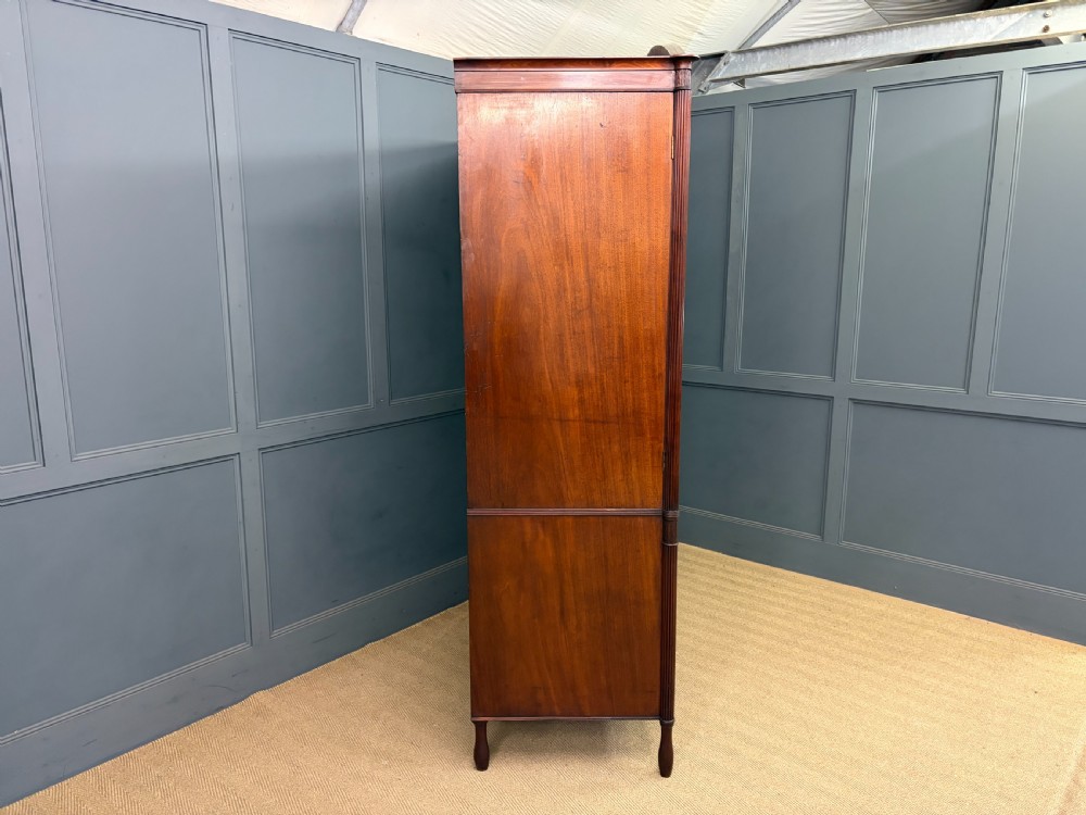 An antique Georgian flame mahogany linen press with an arched top cornice, two inlaid cupboard doors, fluted side pilasters, and a base of five drawers with turned knobs. View XXIV