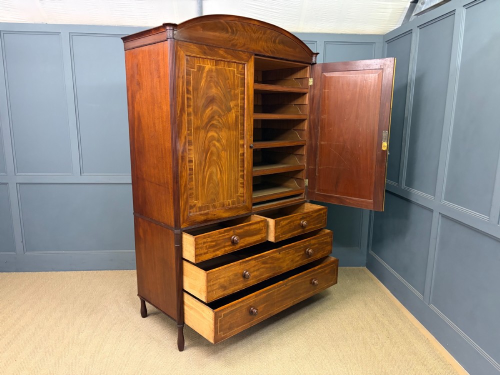 An antique Georgian flame mahogany linen press with an arched top cornice, two inlaid cupboard doors, fluted side pilasters, and a base of five drawers with turned knobs. View XXV