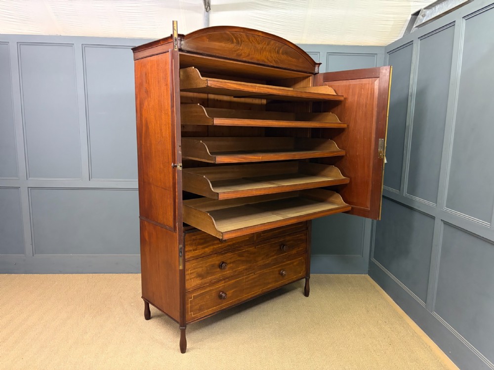 An antique Georgian flame mahogany linen press with an arched top cornice, two inlaid cupboard doors, fluted side pilasters, and a base of five drawers with turned knobs. View XXVI