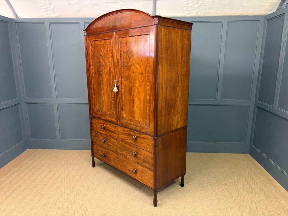 An antique Georgian flame mahogany linen press with an arched top cornice, two inlaid cupboard doors, fluted side pilasters, and a base of five drawers with turned knobs. View XXVII