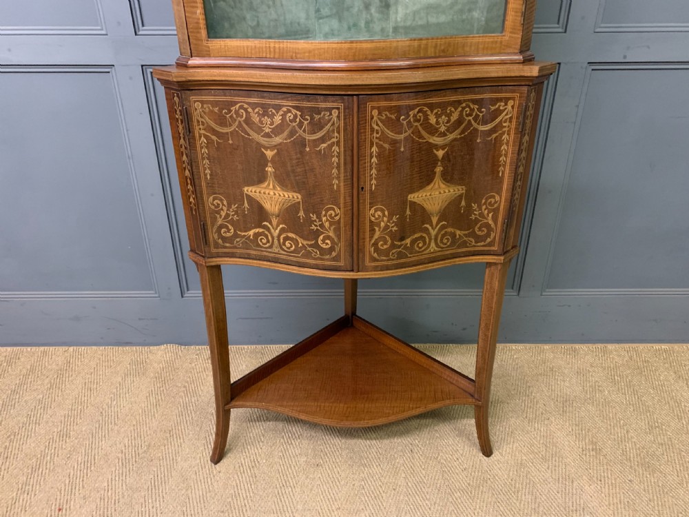 An Edwardian inlaid mahogany corner display cabinet featuring a swan-neck pediment, a glass door with a green fabric lining, neoclassical urn marquetry on the lower doors, and slender legs with an undertier. View II