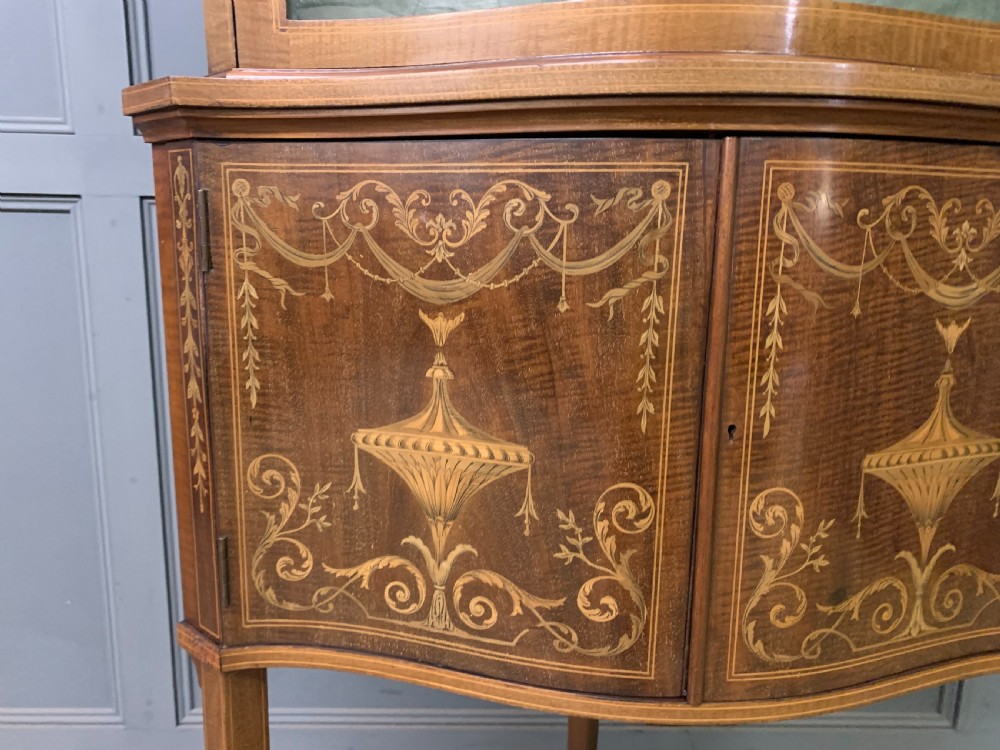 An Edwardian inlaid mahogany corner display cabinet featuring a swan-neck pediment, a glass door with a green fabric lining, neoclassical urn marquetry on the lower doors, and slender legs with an undertier. View III