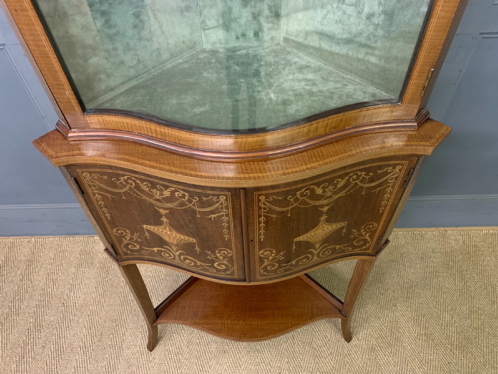 An Edwardian inlaid mahogany corner display cabinet featuring a swan-neck pediment, a glass door with a green fabric lining, neoclassical urn marquetry on the lower doors, and slender legs with an undertier. View IV