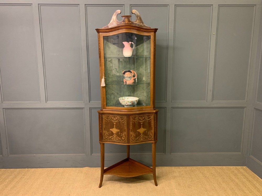 An Edwardian inlaid mahogany corner display cabinet featuring a swan-neck pediment, a glass door with a green fabric lining, neoclassical urn marquetry on the lower doors, and slender legs with an undertier. View VI
