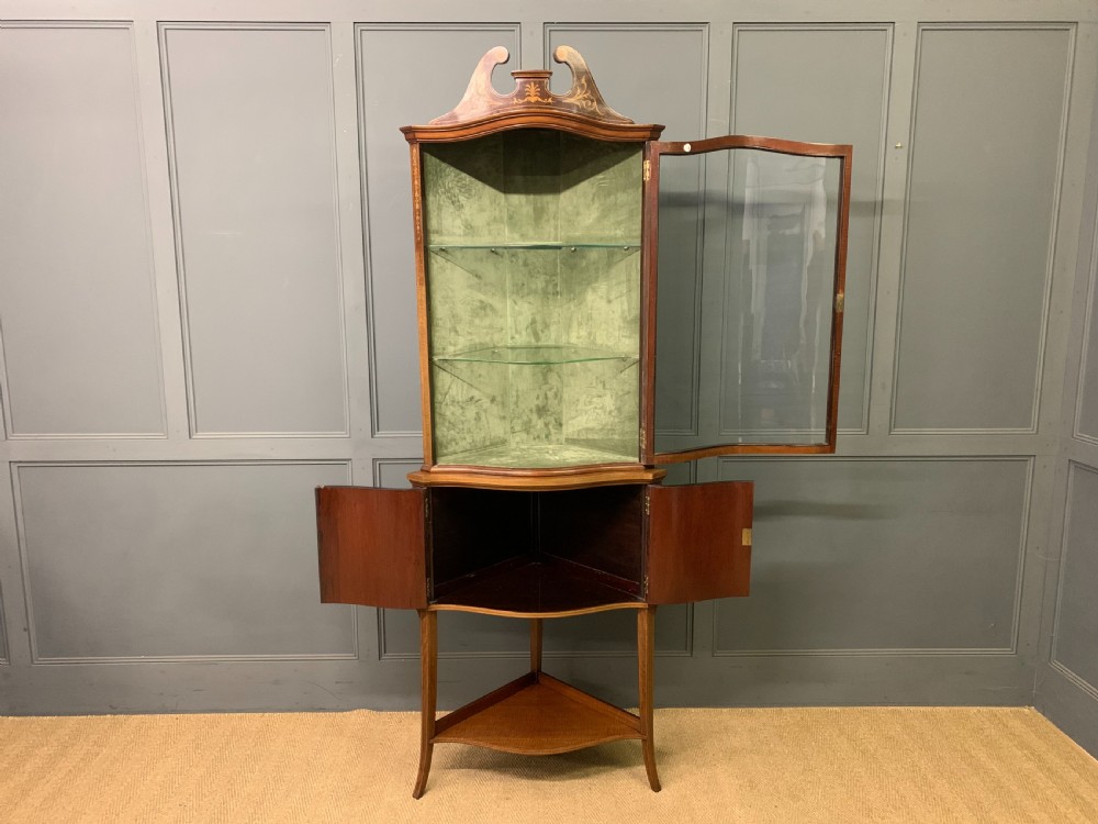 An Edwardian inlaid mahogany corner display cabinet featuring a swan-neck pediment, a glass door with a green fabric lining, neoclassical urn marquetry on the lower doors, and slender legs with an undertier. View VII