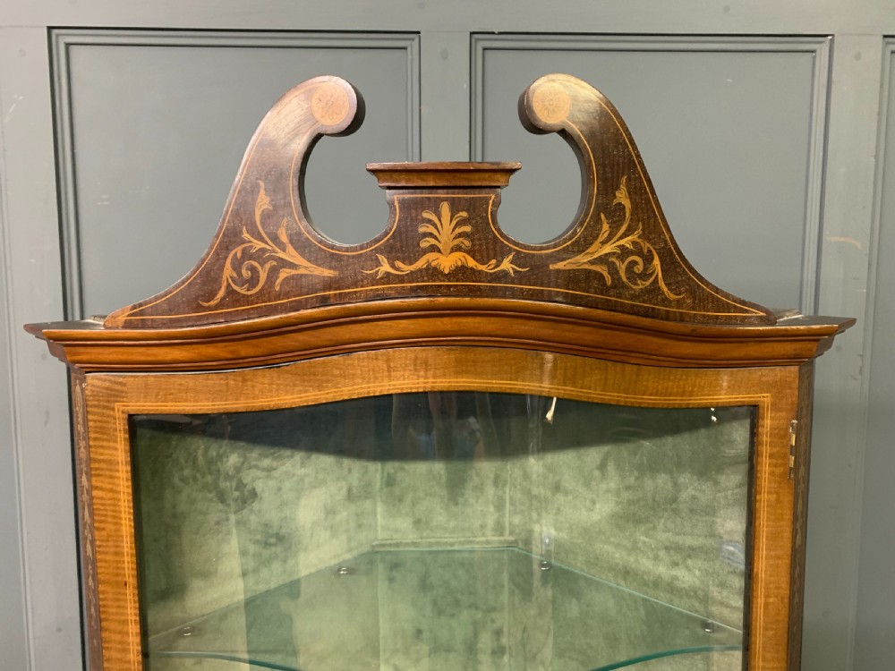 An Edwardian inlaid mahogany corner display cabinet featuring a swan-neck pediment, a glass door with a green fabric lining, neoclassical urn marquetry on the lower doors, and slender legs with an undertier. View VIII
