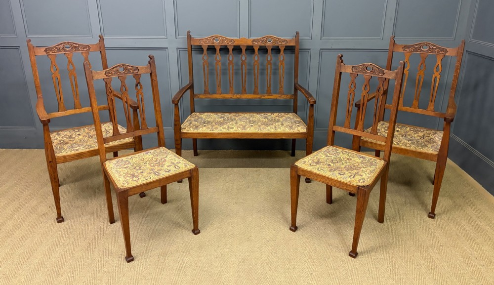 A five-piece Arts and Crafts oak salon suite comprising a two-seater settee, two armchairs, and two side chairs with carved crest rails and patterned upholstery.