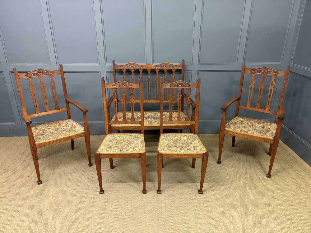 A five-piece Arts and Crafts oak salon suite comprising a two-seater settee, two armchairs, and two side chairs with carved crest rails and patterned upholstery. View II