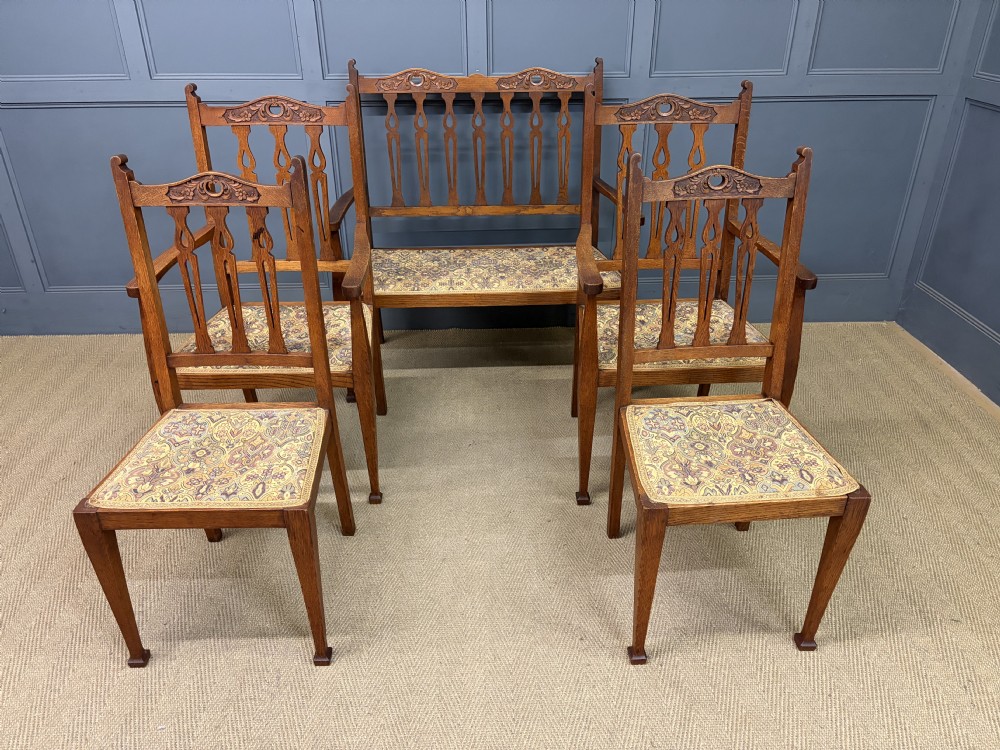 A five-piece Arts and Crafts oak salon suite comprising a two-seater settee, two armchairs, and two side chairs with carved crest rails and patterned upholstery. View III