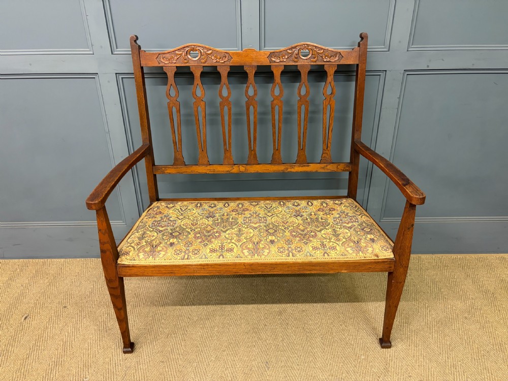 A five-piece Arts and Crafts oak salon suite comprising a two-seater settee, two armchairs, and two side chairs with carved crest rails and patterned upholstery. View IV