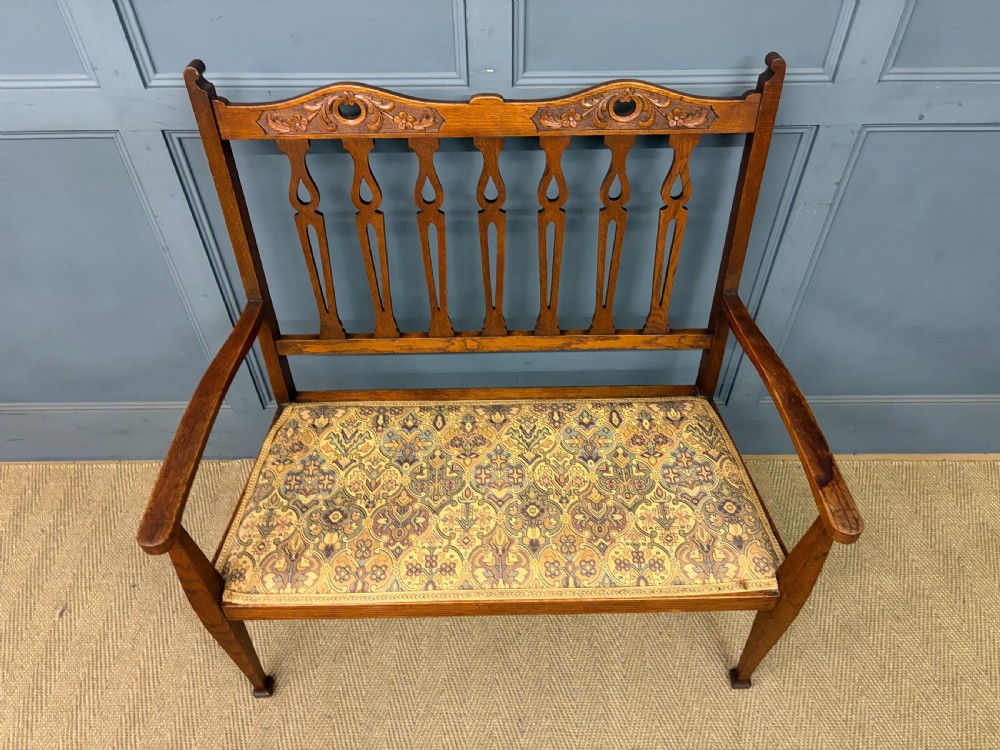 A five-piece Arts and Crafts oak salon suite comprising a two-seater settee, two armchairs, and two side chairs with carved crest rails and patterned upholstery. View V