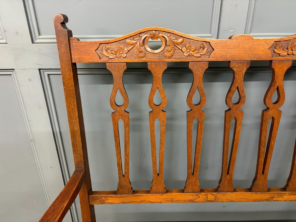 A five-piece Arts and Crafts oak salon suite comprising a two-seater settee, two armchairs, and two side chairs with carved crest rails and patterned upholstery. View VI