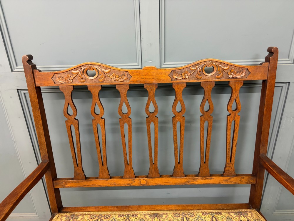 A five-piece Arts and Crafts oak salon suite comprising a two-seater settee, two armchairs, and two side chairs with carved crest rails and patterned upholstery. View VII