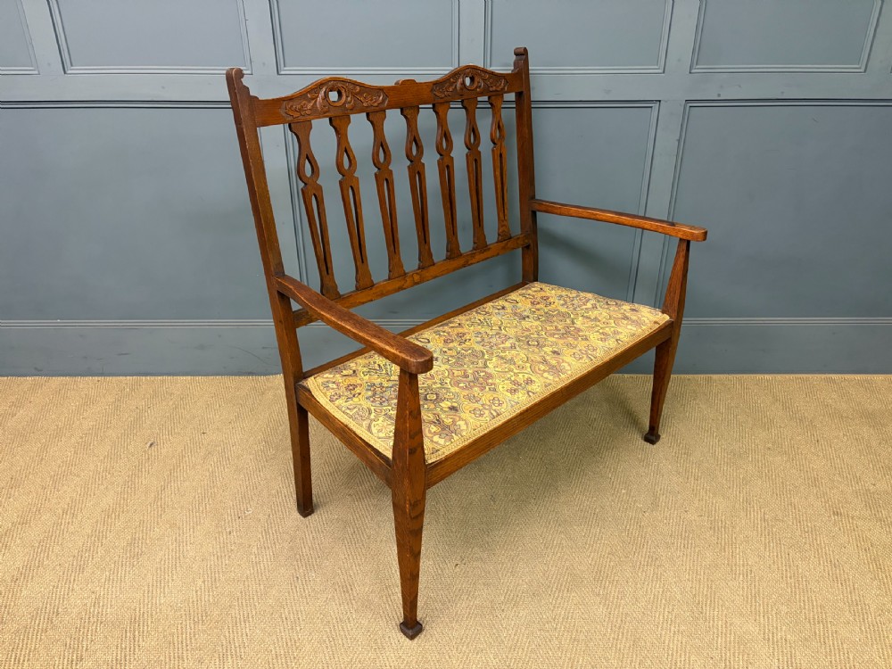A five-piece Arts and Crafts oak salon suite comprising a two-seater settee, two armchairs, and two side chairs with carved crest rails and patterned upholstery. View IX