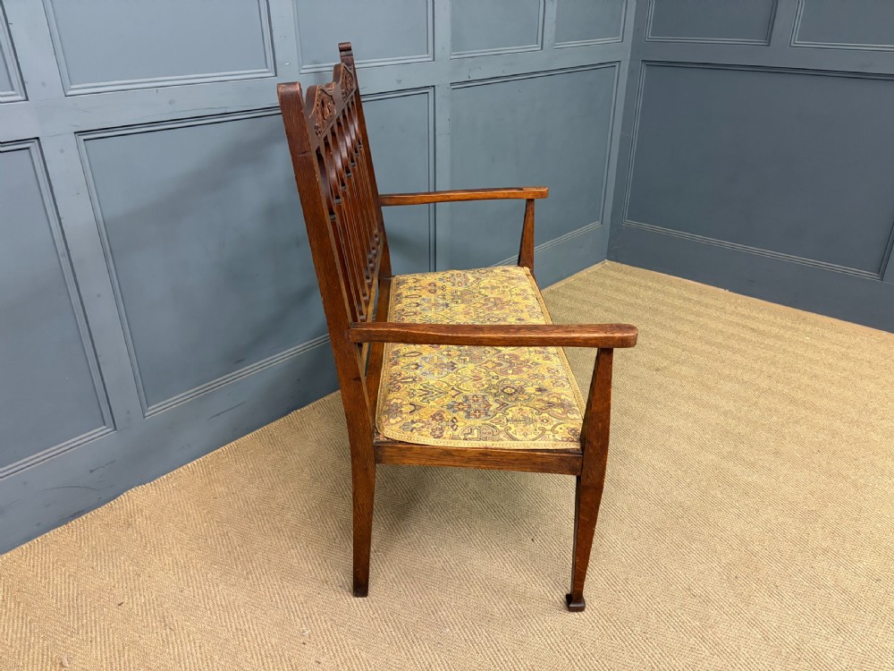 A five-piece Arts and Crafts oak salon suite comprising a two-seater settee, two armchairs, and two side chairs with carved crest rails and patterned upholstery. View X
