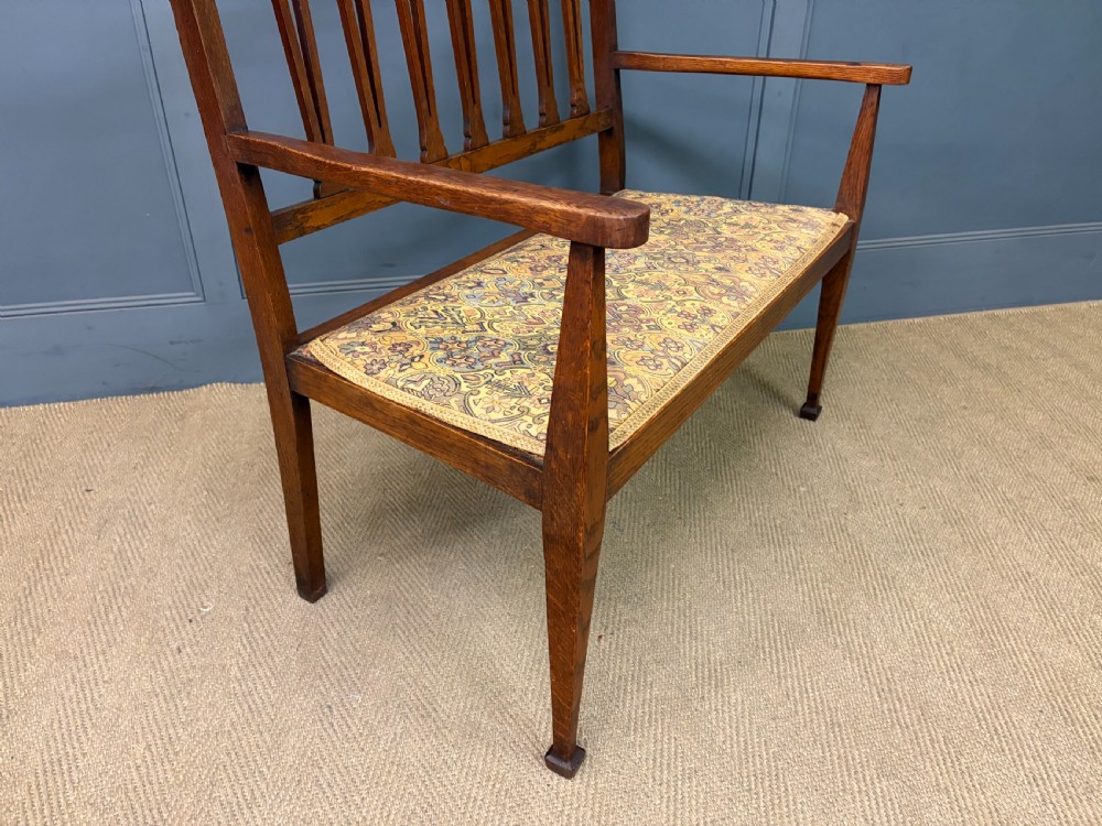 A five-piece Arts and Crafts oak salon suite comprising a two-seater settee, two armchairs, and two side chairs with carved crest rails and patterned upholstery. View XI