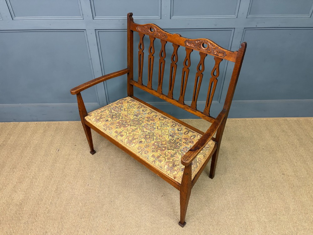 A five-piece Arts and Crafts oak salon suite comprising a two-seater settee, two armchairs, and two side chairs with carved crest rails and patterned upholstery. View XII