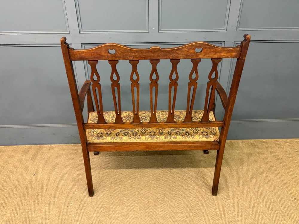 A five-piece Arts and Crafts oak salon suite comprising a two-seater settee, two armchairs, and two side chairs with carved crest rails and patterned upholstery. View XIII