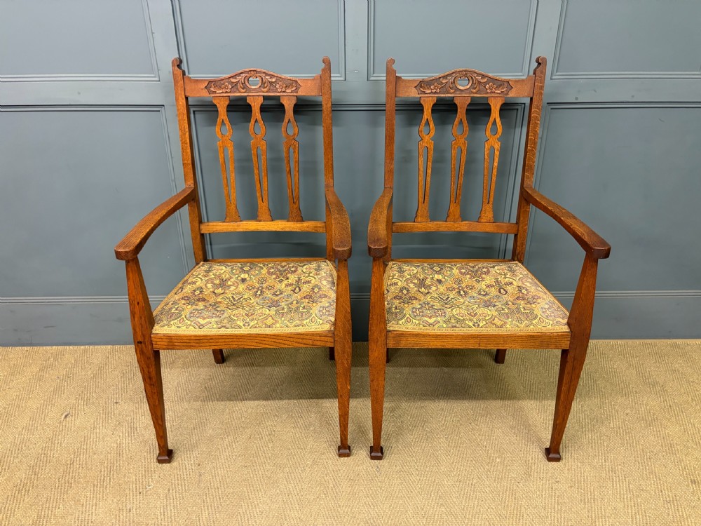 A five-piece Arts and Crafts oak salon suite comprising a two-seater settee, two armchairs, and two side chairs with carved crest rails and patterned upholstery. View XV