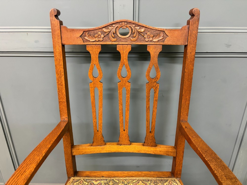 A five-piece Arts and Crafts oak salon suite comprising a two-seater settee, two armchairs, and two side chairs with carved crest rails and patterned upholstery. View XIX