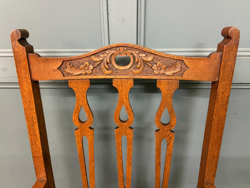 A five-piece Arts and Crafts oak salon suite comprising a two-seater settee, two armchairs, and two side chairs with carved crest rails and patterned upholstery. View XX