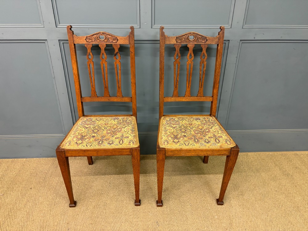 A five-piece Arts and Crafts oak salon suite comprising a two-seater settee, two armchairs, and two side chairs with carved crest rails and patterned upholstery. View XXV