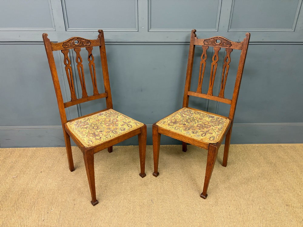 A five-piece Arts and Crafts oak salon suite comprising a two-seater settee, two armchairs, and two side chairs with carved crest rails and patterned upholstery. View XXVI