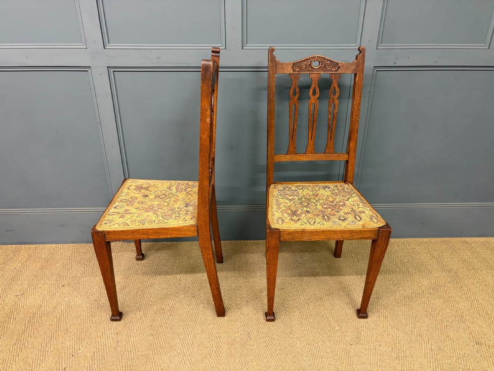 A five-piece Arts and Crafts oak salon suite comprising a two-seater settee, two armchairs, and two side chairs with carved crest rails and patterned upholstery. View XXVII