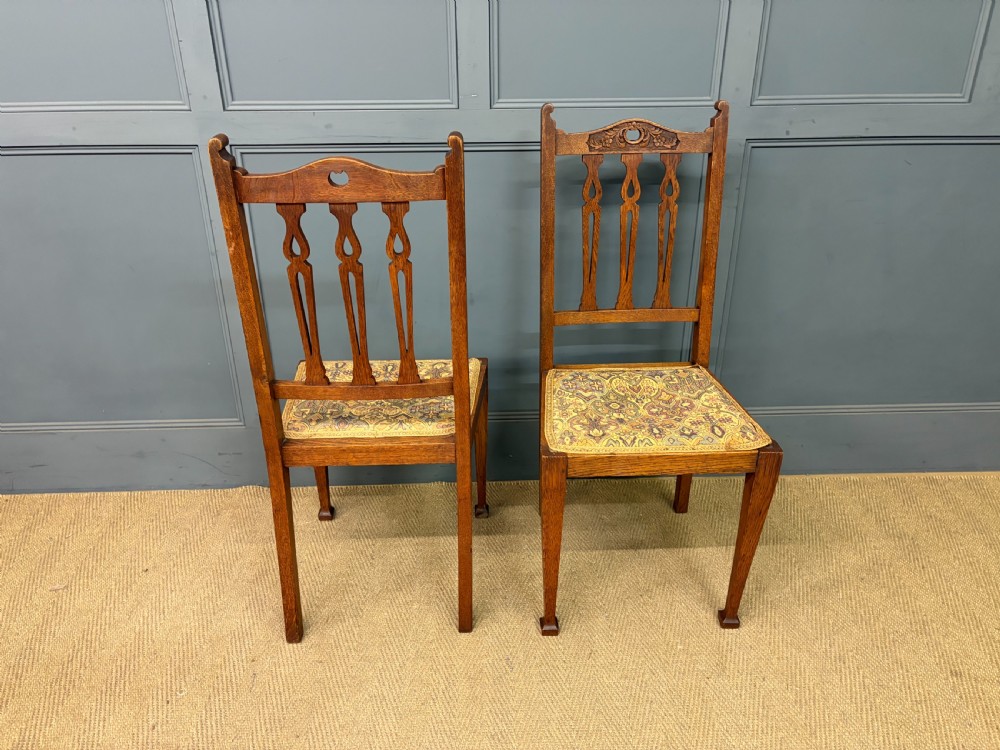 A five-piece Arts and Crafts oak salon suite comprising a two-seater settee, two armchairs, and two side chairs with carved crest rails and patterned upholstery. View XXVIII