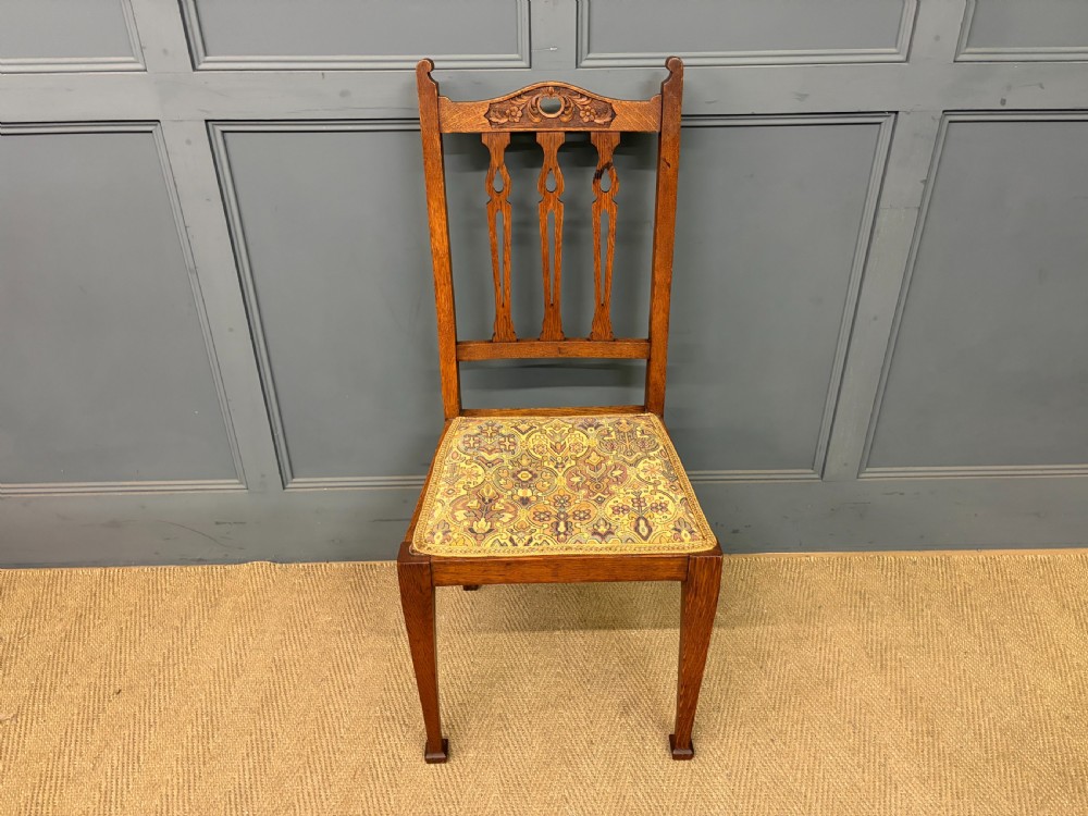 A five-piece Arts and Crafts oak salon suite comprising a two-seater settee, two armchairs, and two side chairs with carved crest rails and patterned upholstery. View XXIX