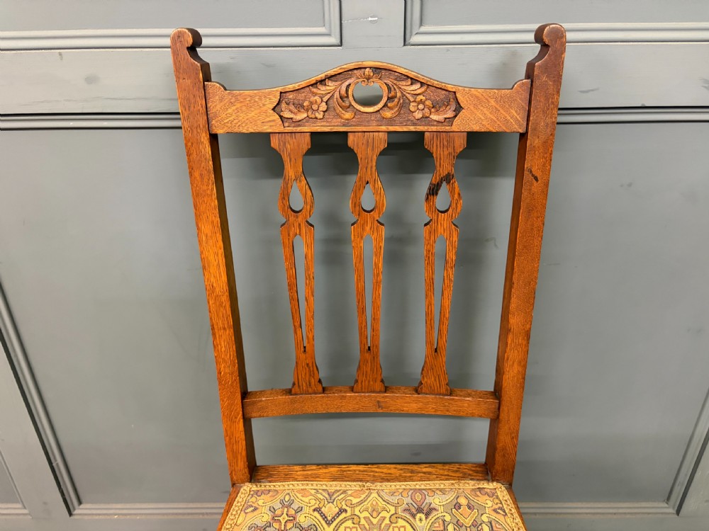 A five-piece Arts and Crafts oak salon suite comprising a two-seater settee, two armchairs, and two side chairs with carved crest rails and patterned upholstery. View XXX