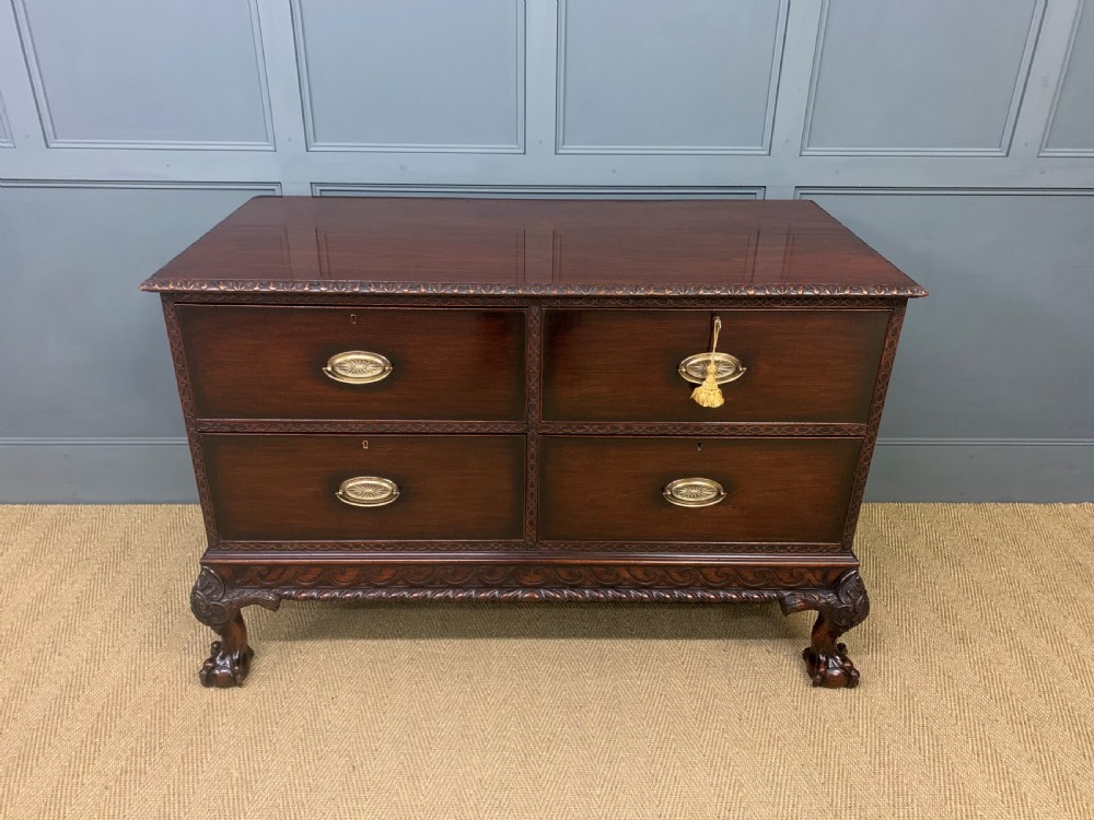 A wide dark mahogany Chippendale style chest featuring four drawers with oval brass handles, carved frieze detailing, and carved ball-and-claw feet.