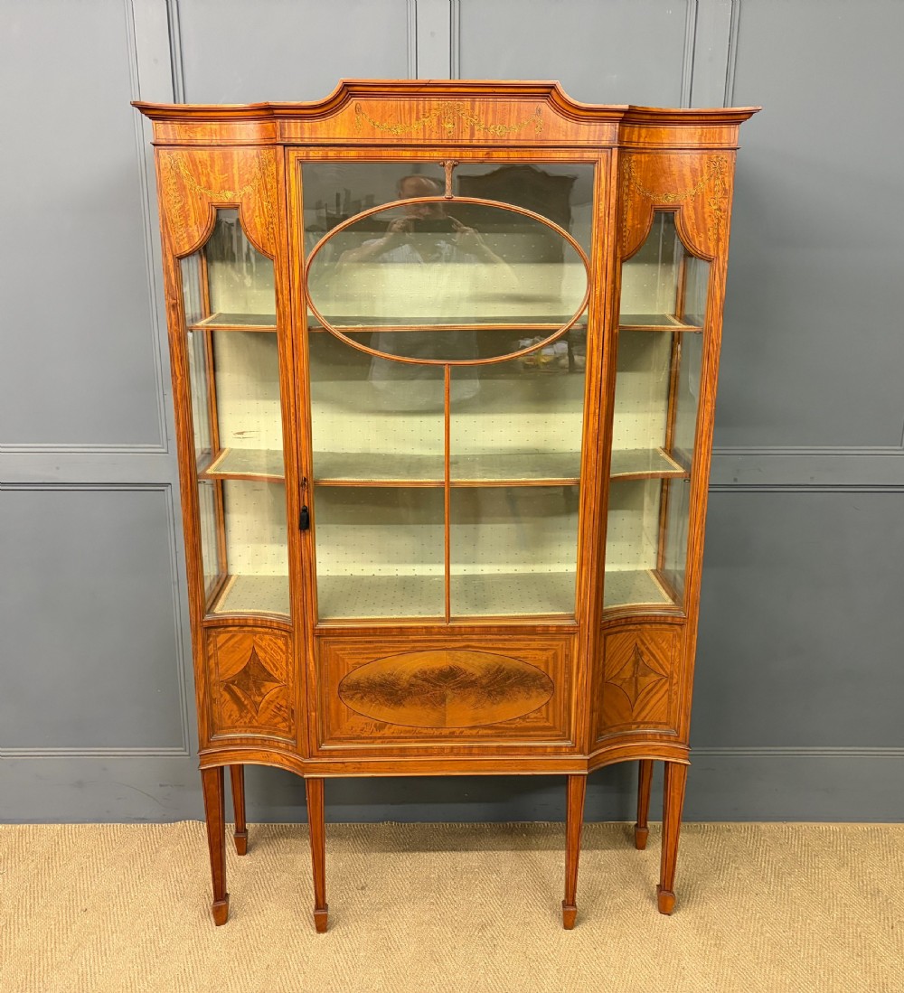 An Edwardian inlaid satinwood display cabinet featuring a central glazed door with an oval wood frame, fabric-lined shelves, and tapered square legs.