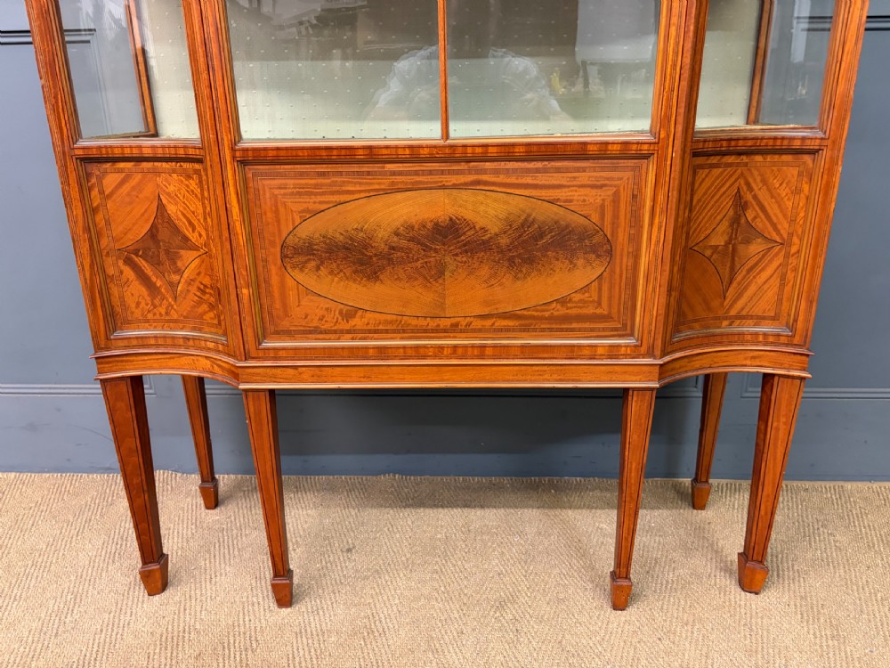 An Edwardian inlaid satinwood display cabinet featuring a central glazed door with an oval wood frame, fabric-lined shelves, and tapered square legs. View IV