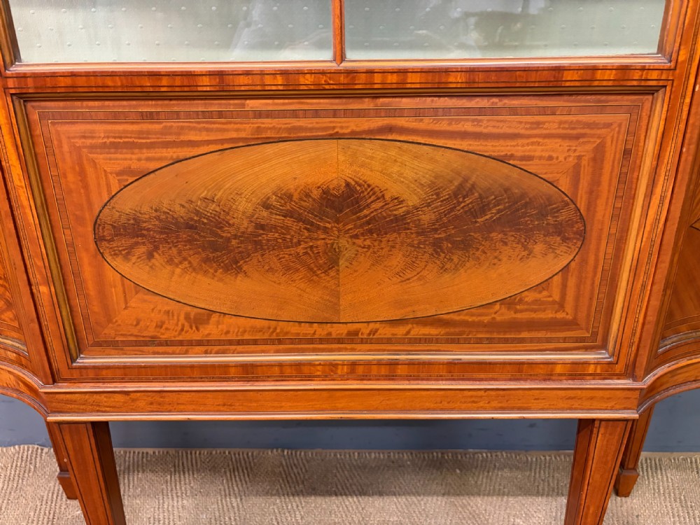 An Edwardian inlaid satinwood display cabinet featuring a central glazed door with an oval wood frame, fabric-lined shelves, and tapered square legs. View V